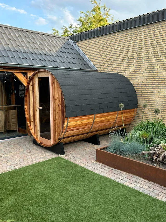 Houten barrelsauna met donkere dakbekleding naast een woning.